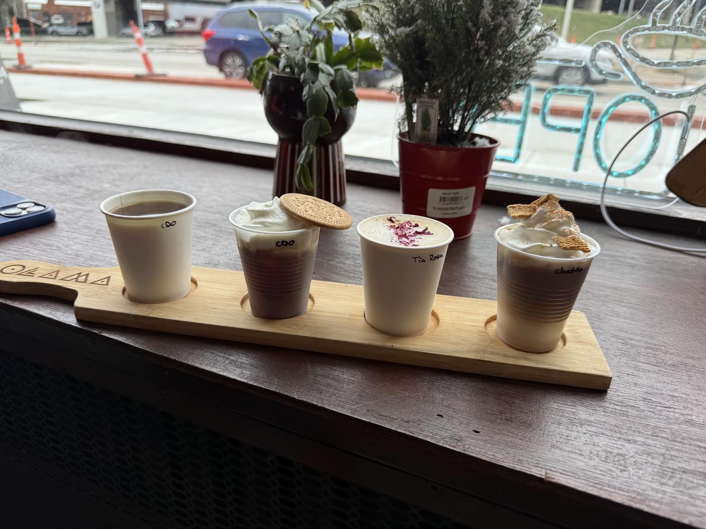 A flight at Café Ollama: four cups on a bamboo paddle engraved Ollama, each cup labelled by hand in marker.